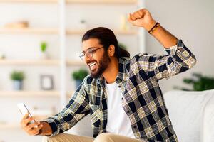 Emotional long-haired indian guy with glasses sitting on couch, using smartphone, raising hand up, happy millennial arab man chilling at home, gambling or trading, side view, copy space photo
