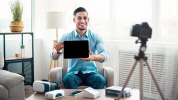 Arab guy influencer shooting review of modern tablet computer on professional dslr camera at home, mockup for design. Cheerful blogger advertising innovative tech product, streaming live on web photo
