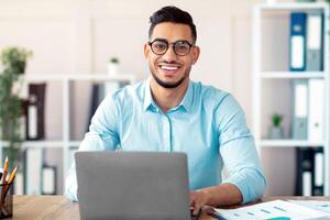 Happy young Arab businessman in glasses sitting at desk, working on laptop at company office. Successful smiling CEO having online job, typing on pc keyboard, having call photo