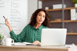 Elearning Concept. Portrait of serious lady teacher having conference call using pc. Lady in wireless headset talking to webcam, shooting tutorial on computer for students pointing at board photo