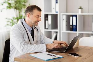 Healthcare And Telemedicine Concept. Portrait of smiling middle aged general practitioner using computer laptop, typing on keyboard, writing report, sitting at desk in his office at clinic photo
