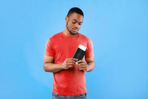 Upset African American Tourist Guy Holding Tickets After Flight Cancellation Standing In Studio On Blue Background. Unhappy Traveler Male Posing With Boarding Pass And Passport Having Problems photo