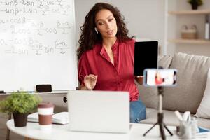 Lady math teacher making conference chat with students using laptop and cellphone on tripod, holding and showing tablet with empty screen with copy space for mockup, explaining photo