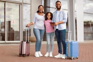Portrait Of Cheerful African American Family Of Three People Standing Outdoors Near Modern Airport Terminal With Suitcases, Dad Holding Passports And Tickets, Looking At Camera And Posing photo