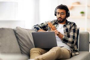 Emotional indian guy sitting on couch at home, having call with partner, yelling and gesturing at notebook screen, using headset, middle-eastern man fighting with colleagues or lover photo