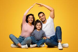 Family Insurance Concept. Happy arab parents making symbolic roof of hands above little daughter, middle eastern mom and dad sitting together with child on floor over yellow background in studio photo