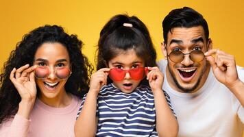 Amazing Offer. Portrait Of Excited Arab Family Of Three Wearing Colorful Sunglasses Looking At Camera With Amazement, Parents And Little Daughter Opening Mouth With Excitement, Yellow Background photo