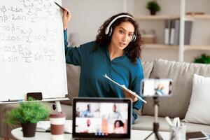 E-Learning, Online Life. Confident tutor in wireless headphones having videocall conference with students and class group using pc, recording vlog with smartphone on tripod due to quarantine lockdown photo