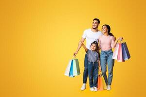 Shopping Offer. Smiling Arab Family With Little Daughter Carrying Shopper Bags And Pointing Aside At Copy Space Over Yellow Background, Happy Parents And Female Kid Enjoying Seasonal Sales, Panorama photo