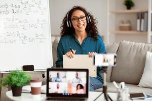 Studying Online. Smiling lady teacher in eyeglasses and headphones giving lesson on web, using smartphone and pc. Millennial lecturer having call with students recording vlog photo