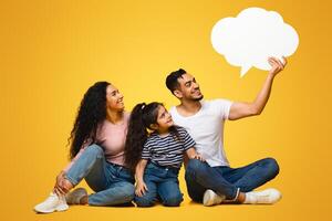 Nice Idea. Happy Arab Family With Little Daughter Looking At Empty Speech Bubble In Father's Hands, Middle Eastern Parents And Child Excited About Interesting Offer, Yellow Background, Copy Space photo