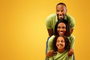 Promo concept. Happy black family of three looking aside at free copy space on yellow studio background, standing behind each other. Empty space for dvertisement photo