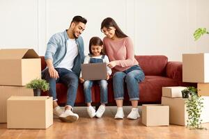 Happy Loving Family Using Laptop Sitting On Couch After Moving New Home. Real Estate, House Ownership And Relocation Concept. People Browsing Internet Sitting Among Carton Boxes, Pointing At Screen photo