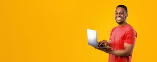 Freelance Career. Happy Black Guy Using Laptop Computer Smiling To Camera Standing Over Yellow Background, Studio Shot. People And Gadgets, E-Learning And Internet Technologies Concept photo