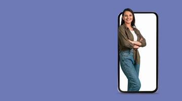 A woman stands confidently while leaning against a large smartphone screen. The person appears engaged, showcasing modern social media usage in a relaxed setting with a plain background. photo