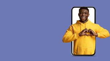 A young man wearing a yellow hoodie smiles as he creates a heart shape with his hands while interacting with a large smartphone screen. The background is a solid color, enhancing the focus on him. photo