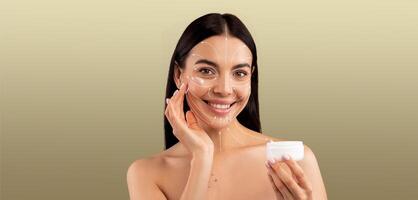 A woman enjoys a skincare routine, applying cream to her face with a joyful smile. She is isolated against a soft, neutral background, creating a peaceful atmosphere for pampering. photo