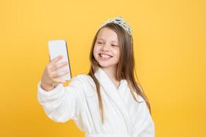 A cheerful girl in a white robe and crown smiles while taking a selfie. The bright yellow background enhances her joyful expression, capturing a moment of fun and childhood innocence. photo