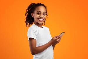 Portrait of smiling African American girl holding smartphone and looking at camera, standing isolated on yellow studio background. Happy black kid chatting online, typing sms, using app, banner photo