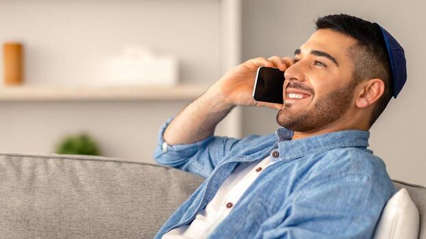 móvil comunicación concepto. perfil lado ver retrato de sonriente joven judío chico en kipa hablando en teléfono inteligente, trabajando remotamente acostado propensión en cómodo sofá con ordenador portátil a hogar oficina foto