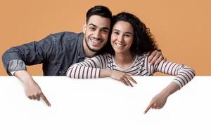 Happy Young Arab Couple Pointing Down At Big White Advertisement Board, Smiling Middle Eastern Spouses Showing Free Copy Space For Your Ad Or Design While Posing Over Yellow Studio Background photo