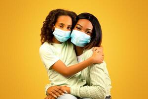 Portrait of african american mother and daughter wearing protective medical masks on face and looking at camera over yellow background. Black mom and her child following measures against coronavirus photo