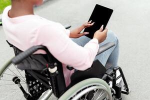 Young black impaired woman in wheelchair using tablet computer with empty screen at autumn park, mockup for app or website. Disabled African American lady with touch pad outdoors, space for template photo