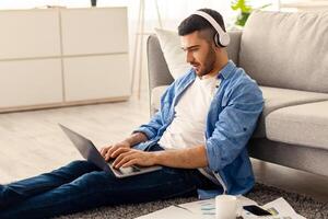 Focused jewish freelancer guy working remotely with laptop at home office, using computer for online project or education, typing on keyboard, sitting on floor leaning on couch, free copy space photo