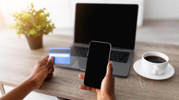 Closeup of young black lady with smartphone, laptop and credit card shopping online, using new mobile app or website to order goods on internet from home during covid quarantine, mockup for design photo