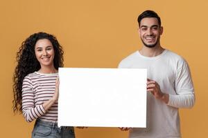 Ad Mockup. Happy Young Arab Spouses Holding White Blank Placard In Hands, Smiling Middle Eastern Couple Demonstrating Copy Space For Your Design While Standing Over Yellow Studio Background photo