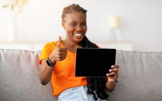 Positive Afro woman showing digital tablet with mockup for shopping app or website, showing thumb up at home, selective focus. Happy black lady buying gifts on web. Internet purchases concept photo