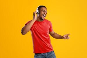 Funny African Man Singing And Dancing Having Fun Posing Wearing Wireless Earphones In Studio Over Yellow Background. New Musical Application Advertisement. Black Guy Enjoying Great Playlist photo