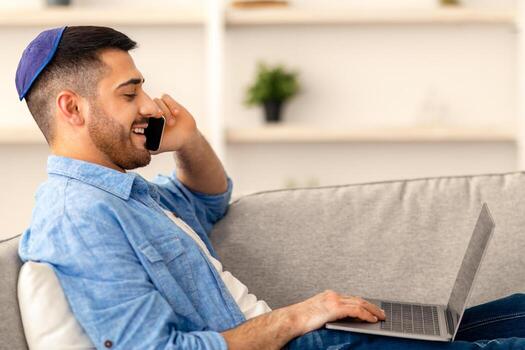 teletrabajo concepto. perfil lado ver retrato de judío empresario hablando en Teléfono móvil utilizando ordenador portátil a hogar lugar de trabajo, sentado en sofá. gerente Discutiendo acuerdo con socios, consultante cliente foto