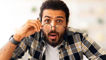 Excited arab guy freelancer looking at camera and pulling eyeglasses down, having amazed face expression, using computer keyboard, middle-eastern man working from home, closeup portrait photo