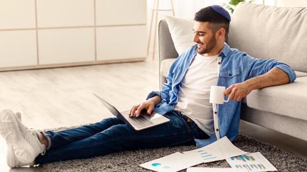 distante trabajo y Lanza libre carrera concepto. sonriente milenario judío chico trabajando remotamente desde hogar sentado en piso alfombra en vivo habitación, Bebiendo café. hombre utilizando ordenador portátil haciendo papeleo tarea foto