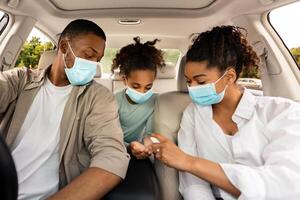 Black Family Spraying Sanitizer On Hands In Car. Parents And Daughter Traveling By Automobile, Wearing Face Masks And Disinfecting Hands For Covid-19 Protection photo