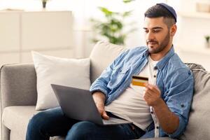 Electronic Payment Service Concept. Portrait of casual young jewish businessman in kippa holding credit card and using laptop, making e-payments while working with computer at home office, copy space photo