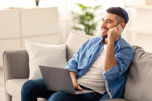 teletrabajo masculino judío empresario hablando en Teléfono móvil utilizando ordenador portátil a hogar lugar de trabajo, sentado en sofá. acuerdo discusión con socios, planificación futuro reuniones, cliente consultante concepto foto
