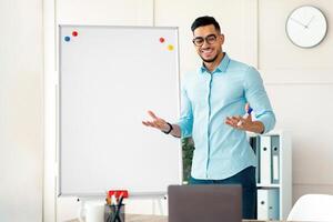 Cheerful young Arab teacher standing near blackboard and explaining English grammar to students online from home. Positive guy tutor explaining foreign language rules on web photo