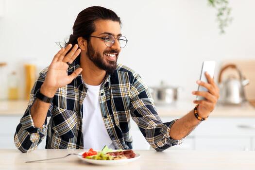 contento joven árabe hombre en casual atuendo teniendo en línea fiesta con familia o amigos vía teléfono inteligente mientras disfrutando su sano desayuno o almuerzo, ondulación a Teléfono móvil pantalla, cocina interior, Copiar espacio foto