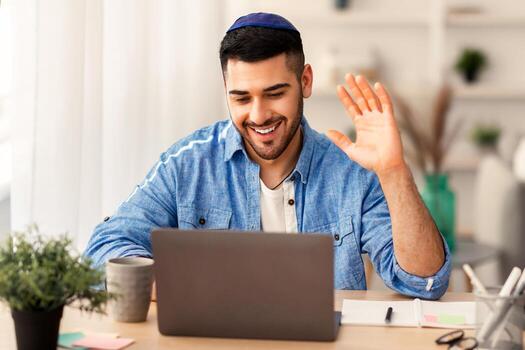 distante comunicación concepto. retrato de contento judío masculino en kippa y casual camisa haciendo llamada sentado a escritorio a hogar oficina y hablando con colegas o familia, ondulación mano a cámara web foto