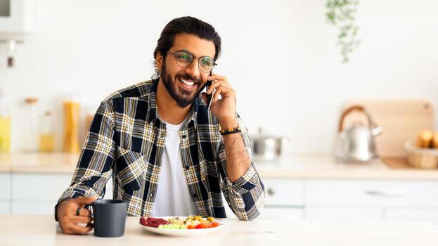hermoso indio chico persona de libre dedicación teniendo teléfono conversacion mientras comiendo a cocina, contento Medio este joven barbado hombre sonriente y mirando a Copiar espacio, comiendo Fresco vegetal ensalada foto