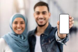Beat App. Cheerful Islamic Couple Showing Smartphone With Blank Black Screen At Camera While Standing In Airport Terminal, Happy Muslim Spouses Recommending New Mobile Application, Mockup Image photo