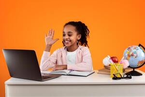 Portrait of smiling African American schoolgirl sitting at desk, making call using personal computer. Cheerful young black pupil raising hand for an answer or to ask a question at virtual class photo