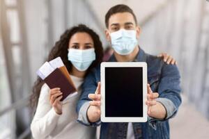 Arab Couple Wearing Medical Face Masks Demonstrating Digital Tablet With Black Blank Screen While Standing In Airport With Passports And Tickets, Spouses Showing Copy Space, Selective Focus, Mockup photo