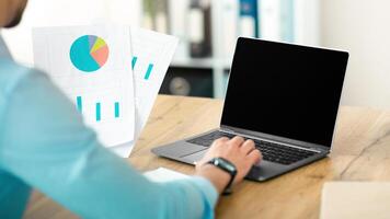 Unrecognizable Arab businessman working on laptop computer with empty screen at company office, mockup for website design. Guy manager with documents and portable pc, space for advertisement photo