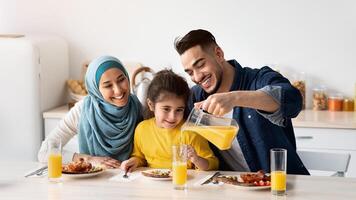 Portrait Of Happy Middle Eastern Family Of Three Having Breakfast In Kitchen Together, Arab Parents With Cute Little Daughter Enjoying Tasty Food And Drinking Orange Juice While Sitting At Table photo