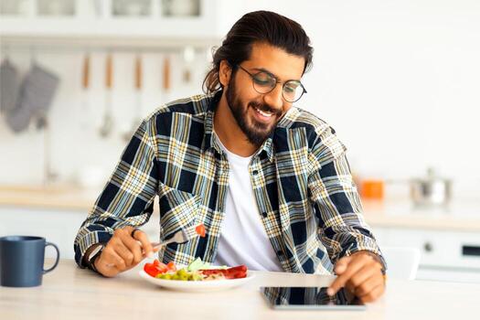 positivo pelo largo, barbado árabe chico teniendo desayuno, comiendo Fresco verduras, salchichas, Bebiendo café y utilizando moderno digital tableta, acecho serie o videos en línea, cocina interior foto