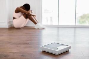 Weight Loss Failure. Unhappy Sporty Lady Crying Sitting In Corner After Weighing Herself And Gaining Kilograms. Selective Focus On Scales, Blurred Background, Shallow Depth. Excess Problem Concept photo