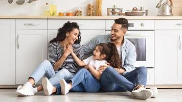 Beautiful middle eastern family father, mother and daughter sitting on floor at cozy white kitchen, loving parents enjoying time with their cute little child, having conversation, laughing, copy space photo
